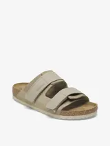 Sandalia Birkenstock modelo Uji en color taupe, confeccionada con una parte superior de gamuza suave y correas de nobuk con cierre de velcro. Presenta la clásica plantilla anatómica de corcho y suela de EVA.