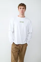 Remera de manga larga color blanco con cuello redondo y diseño de doble capa en las mangas. Presenta un estampado pequeño en el pecho con la frase 'Napoli 1990 Ciao, ci vediamo' en color verde.