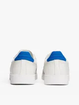 Zapatillas deportivas de estilo vintage Tommy Jeans modelo The Greenwich, confeccionadas en ante color blanco con paneles laterales en azul vibrante y detalles en gamuza gris claro. Presentan cierre con cordones, suela cupsole de goma y logo de la marca en el lateral.
