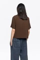 Camiseta de manga corta y escote redondo, color beige claro, con corte boxy. Presenta una hilera de tachuelas metálicas negras decorativas en la parte superior del pecho y en las costuras de los hombros.