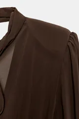 Vestido corto de color marrón con diseño combinado. Presenta un cuerpo superior fluido con cuello subido, lazada frontal ajustable y mangas largas abullonadas con hombreras y puños ajustados. La parte inferior es entallada con tejido elástico a tono, espalda con escote fluido y cierre lateral con cremallera oculta.