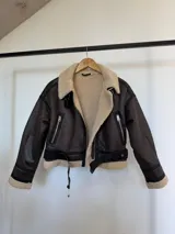 Chaqueta tipo gamulán de cuero sintético en color marrón oscuro, con interior y solapas de corderito sintético en tono crema. Presenta cierre frontal, bolsillos laterales con cierre, correas ajustables en la cintura y detalles de terminación en los puños.