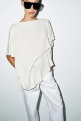 Top tipo capa blanco de punto fluido, con mezcla de viscosa, cuello bote, mangas cortas y bajo asimétrico.