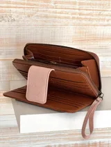 Billetera grande de cuero color marrón con solapa rosa y cierre de cremallera. Incluye correa de mano extraíble.