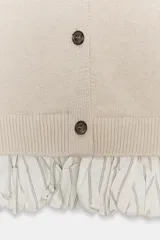 Chaqueta de punto color beige con cuello redondo, manga larga y cierre frontal con botones. Presenta un diseño combinado con volados de tela en el bajo.