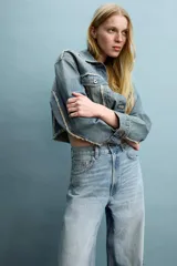 Chaqueta de jean estilo crop, con cuello solapa, manga larga y cierre frontal con botones metálicos. Presenta bolsillos de plastrón con solapa en el pecho y detalles de costuras deshilachadas en el frente.