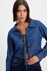 Camisa crop de denim azul con cuello camisero y cierre frontal mediante cordones ajustables a través de ojales. Presenta mangas largas con puños abotonados.