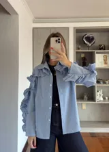 Camisa de corte recto con estampado de cuadros vichy en azul y blanco. Presenta cuello clásico, cierre frontal con botones y detalle de volados decorativos que recorren las mangas desde el hombro hasta el puño.