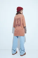 Remera de corte oversize color marrón con efecto gastado, manga corta y estampa gráfica retro en el frente.