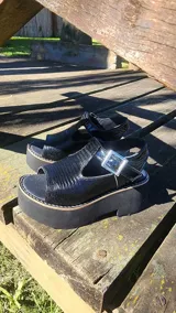Zueco abierto de cuero negro con textura reptil, plataforma alta, tira con hebilla metálica y diseño de horma ancha.