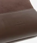 Cartera transversal pequeña color marrón, con la parte superior de gamuza y la inferior de cuero sintético. Tiene cierre de cremallera metálica dorada y asa larga ajustable y desmontable con mosquetones dorados.