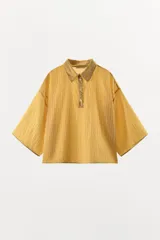 Camisa de corte holgado confeccionada en mezcla de lyocell, con estampado de rayas verticales en tonos amarillos y blancos. Presenta cuello solapa, cierre frontal parcial con botones y mangas anchas por debajo del codo.