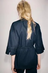 Camisa de denim azul oscuro con cuello solapa, mangas amplias por debajo del codo y bolsillos de parche en el frente. Cuenta con cintura ajustable mediante cordón y cierre frontal con botones metálicos.