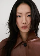 Chaqueta larga de efecto cuero color marrón, con cuello camisero, botonadura frontal oculta, bolsillos laterales y cinturón ajustable con hebilla en la cintura. Cuenta con trabillas en las muñecas y diseño amplio.