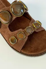 Sandalias planas de gamuza marrón, con dos tiras anchas adornadas con grandes piedras redondas facetadas en tonos marrones con bordes metálicos dorados.