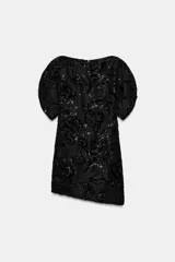 Vestido corto de cuello barco y manga corta abullonada, confeccionado en tejido negro con bordados florales y aplicaciones de abalorios y brillos a tono. Presenta un diseño con volumen en las mangas y cierre de cremallera oculta en la espalda.