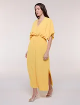 Vestido largo amarillo de corte fluido con escote en V, mangas cortas amplias y apliques de tachas en el escote y puños. Posee cintura elastizada fruncida y falda larga con tajos laterales en el ruedo.