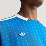 Camiseta de fútbol Adidas del Real Madrid, inspirada en los años 80. Diseño de color azul con finas rayas verticales blancas, cuello en V blanco, mangas cortas con las tres tiras características de la marca y escudo retro del club bordado en el pecho.