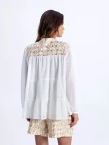 Camisa blanca vaporosa con cuello mao, botonadura frontal oculta y mangas largas abullonadas con puños elásticos. Presenta un diseño de paneles fruncidos y detalles bordados con motivos geométricos dorados en la parte superior delantera.