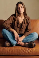 Blusa de color marrón con acabado satinado, cuello en V con volados y lazo de ajuste frontal. Presenta mangas largas con puños elásticos y un corte holgado.