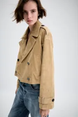 Trench corto cruzado de piel de ante color tostado, con cuello de solapa, bolsillos delanteros de vivo y manga larga con trabillas en los puños.