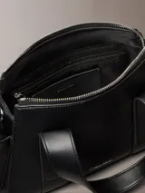Cartera estructurada Calvin Klein de piel sintética negra, con diseño de asas superiores y bolsillos laterales. Cuenta con cierre de cremallera y logo metálico grabado en la parte delantera.