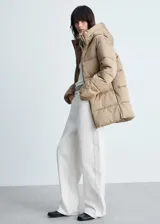 Campera puffer acolchada de color beige, con diseño recto, cuello alto y capucha integrada. Cuenta con cierre frontal de cremallera doble, mangas largas con puños elásticos y dos bolsillos laterales con cierre. Acabado repelente al agua.