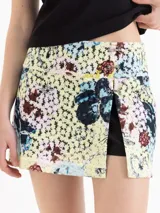 Minifalda de encaje estampado con diseño floral en tonos amarillos, celestes y marrones, que integra un short de poplín negro por debajo. Presenta un tajo frontal y detalles de volados de cuero.