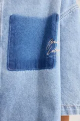 Camisa oversize de denim azul claro, con cuello clásico, mangas cortas a la altura del codo y bolsillo parche en tono más oscuro con detalle bordado en el pecho.
