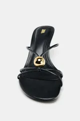 Sandalia de tacón fino color negro, con tiras finas en la parte delantera y detalle de aplique metálico dorado.