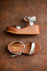 Sandalias de plataforma con suela de madera marrón y capellada de tiras plateadas metálicas. Posee una tira ancha en la parte delantera y una tira ajustable con hebilla en el tobillo. La plantilla es de cuero marrón con el logo de la marca "AGNES".