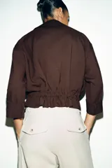 Chaqueta corta marrón de cuello solapa y manga remangada por debajo del codo. Presenta bolsillos delanteros de plastrón con solapa, bajo ajustable con cordón interior y cierre frontal con botones.