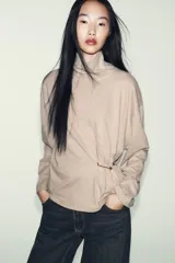 Top de cuello alto y manga larga, confeccionado en tejido de algodón gris melange. Presenta un diseño con frunces laterales ajustados mediante un aplique metálico decorativo.