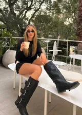 Botas de caña alta confeccionadas en ecocuero negro, con diseño de tres hebillas metálicas laterales y suela plana.
