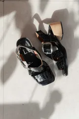 Sandalias de cuero negro con textura de reptil, diseño de punta cuadrada y taco bloque de madera. Presentan tiras cruzadas en el empeine y una hebilla metálica grande decorativa.