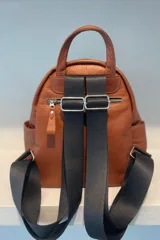 Mochila de cuero gamuzado color marrón con detalles en cuero metalizado plateado. Cuenta con cierre frontal, correas de cinta ajustables y placa metálica con logo de la marca en la parte inferior.