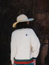 Buzo tejido de punto con escote en V, manga larga y diseño holgado. Presenta un patrón de rayas verticales en relieve y detalles de franjas en contraste en el cuello, puños y ruedo. Incluye un pequeño bordado en el pecho.