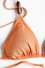 Top de bikini de triángulo color salmón con tiras finas ajustables para atar en la espalda y detalle de cuentas carey en el bretel derecho. Cuenta con copas con relleno extraíble y diseño forrado.