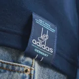 Remera de estilo urbano Adidas inspirada en la selección de fútbol de Alemania. Es de color azul marino con las icónicas tres tiras en celeste sobre los hombros y mangas. Presenta el logo del trébol de Adidas en blanco en el pecho derecho y el escudo de la federación alemana en el izquierdo.