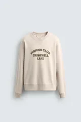 Sudadera de corte relajado color beige, con cuello redondo, manga larga y puños acanalados. Presenta un texto bordado en relieve en el pecho con la leyenda 'Fishing Club Cromwell Lake' en color verde oliva.
