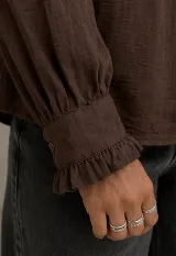Camisa negra de manga larga con detalles de volados en el cuello alto, a lo largo de la botonadura frontal y en los puños elásticos. Presenta frunces en el canesú delantero y cierre frontal con botones.