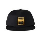 Gorra snapback negra con visera plana y parche frontal rectangular de color dorado con un diseño gráfico abstracto que simula dos caras o formas simétricas.