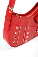 Cartera tipo baguette de color rojo con acabado brillante, decorada con múltiples cierres metálicos, ojales y dos pequeñas hebillas decorativas en el frente. Incluye un bolsillo pequeño con solapa y cierre de botón a presión, además de una correa larga ajustable.