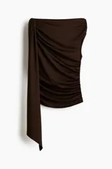 Top sin mangas color marrón oscuro, de punto elástico, con ajuste entallado. Presenta un adorno de metal y una tira larga en un hombro que forma un escote asimétrico. Los laterales están fruncidos para crear un efecto drapeado.