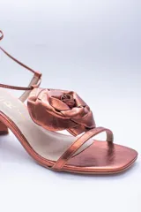Sandalias de cuero color cobre con taco cuadrado y tira fina sobre los dedos. Presentan una flor grande en el empeine y tiras finas para atar al tobillo.