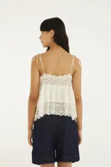 Blusa color blanco off-white, confeccionada en algodón con detalles de encaje floral en el escote y en el ruedo, tirantes finos ajustables con lazo y cierre frontal con pequeños botones.