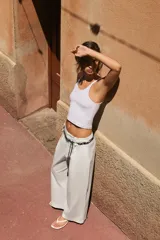 Pantalón de popelina de tiro alto y pernera ancha, de color negro. Presenta pinzas delanteras, bolsillos laterales y falsos bolsillos de vivo en la espalda. Incluye un cinturón combinado tipo pañuelo trenzado con tiras colgantes y cuentas de madera. Cierre frontal con cremallera, botón interior y gancho.