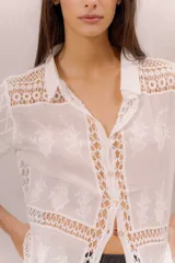 Camisa de algodón color crudo, con cuello camisero, mangas cortas y cierre frontal con botones. Presenta paneles de tejido calado y detalles bordados transparentes.