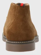 Bota tipo chukka de Hush Puppies, confeccionada en descarne color marrón, con cordones al tono, suela de goma y detalle de tirador en el talón con franjas rojas y negras.