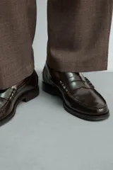 Mocasines de vestir tipo loafer para hombre, confeccionados en cuero marrón oscuro con acabado brillante. Presentan diseño clásico de antifaz en el empeine, costuras visibles en relieve y suela plana con un ligero tacón.